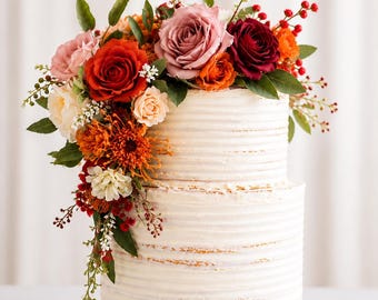 Puede incluir: Un pastel blanco de dos pisos con glaseado texturizado, adornado con un vibrante arreglo floral. Las flores incluyen rosas rojas, naranjas y rosas, con follaje verde y bayas. El pastel se encuentra sobre un soporte blanco.
