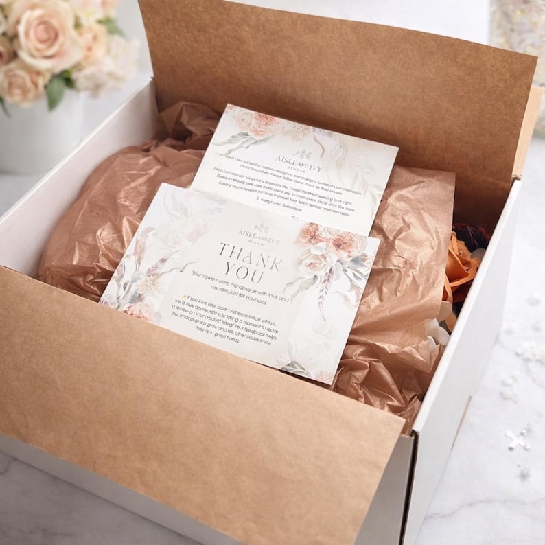 Puede incluir: Una caja de env&iacute;o de cart&oacute;n blanco abierta, forrada con papel de seda marr&oacute;n, que contiene dos tarjetas rectangulares con ilustraciones florales. La tarjeta superior dice Thank You y Aisle and Ivy Studio, con m&aacute;s texto.