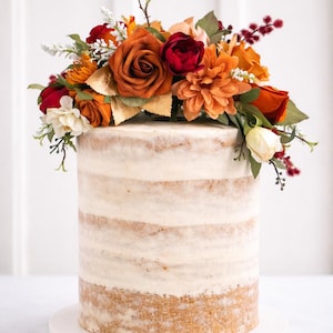 Puede incluir: Un pastel cil&iacute;ndrico alto de estilo semi-naked con capas de bizcocho visibles y glaseado blanco, decorado con un arreglo de flores artificiales en tonos naranja quemado, rojo intenso, crema y follaje verde sobre una base blanca.