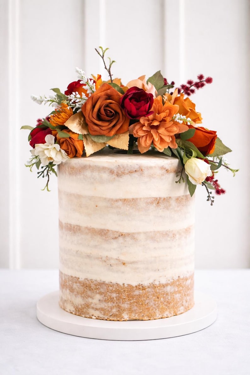Puede incluir: Un pastel cil&iacute;ndrico alto de estilo semi-naked con capas de bizcocho visibles y glaseado blanco, decorado con un arreglo de flores artificiales en tonos naranja quemado, rojo intenso, crema y follaje verde sobre una base blanca.