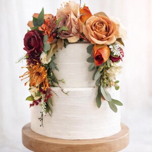 Puede incluir: Un pastel de dos niveles con glaseado blanco decorado con un arreglo de flores artificiales en tonos naranja quemado, burdeos intenso y crema, que incluye rosas y follaje, exhibido sobre un soporte de pedestal de madera redondo.