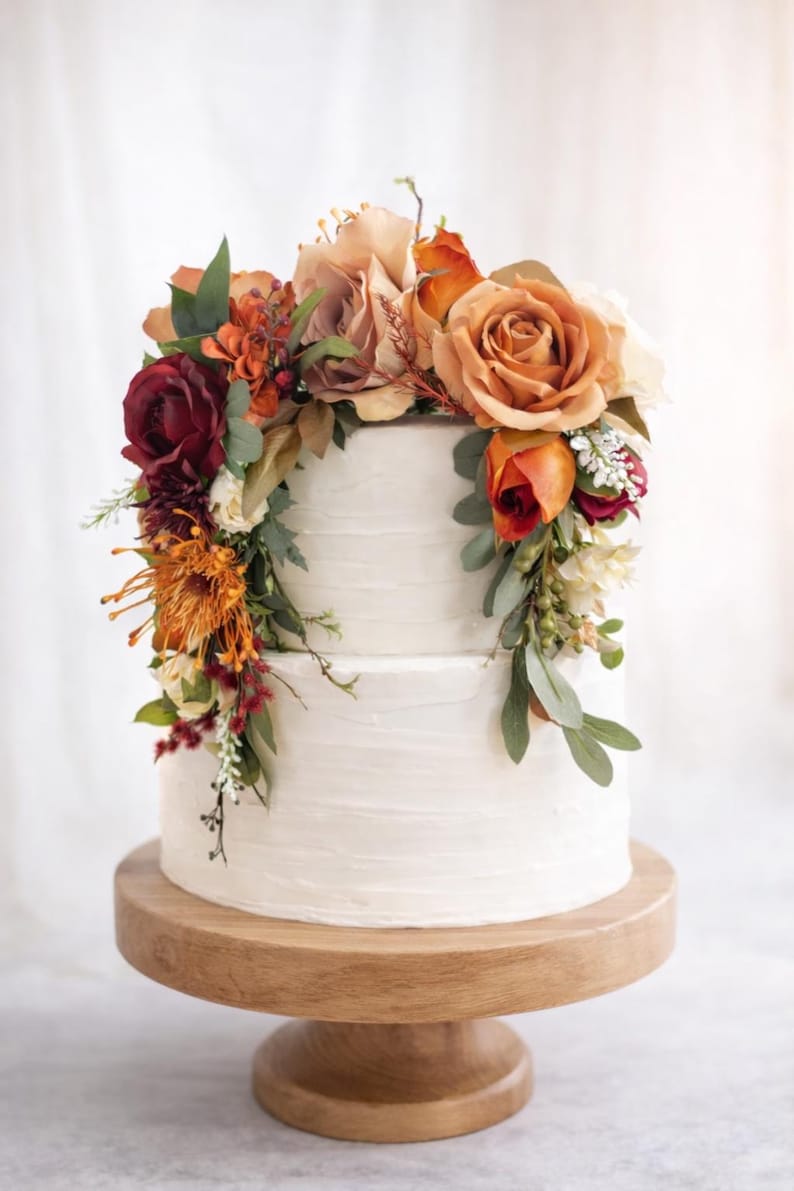 Puede incluir: Un pastel de dos niveles con glaseado blanco decorado con un arreglo de flores artificiales en tonos naranja quemado, burdeos intenso y crema, que incluye rosas y follaje, exhibido sobre un soporte de pedestal de madera redondo.