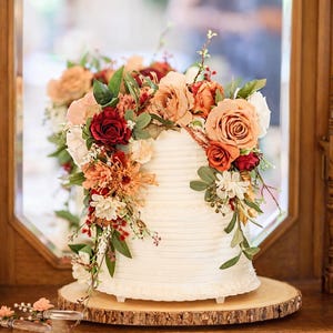 Puede incluir: Un pastel blanco de dos niveles con glaseado de textura horizontal, decorado con un arreglo de rosas artificiales y follaje en tonos rojo intenso, naranja quemado, melocot&oacute;n y crema, sobre una base de madera natural.