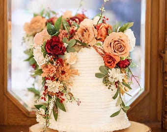 Puede incluir: Una tarta blanca de varios pisos decorada con una cascada de flores rojas, naranjas y melocotón. La tarta se encuentra sobre una rodaja de madera, con un espejo y muebles de madera en el fondo.