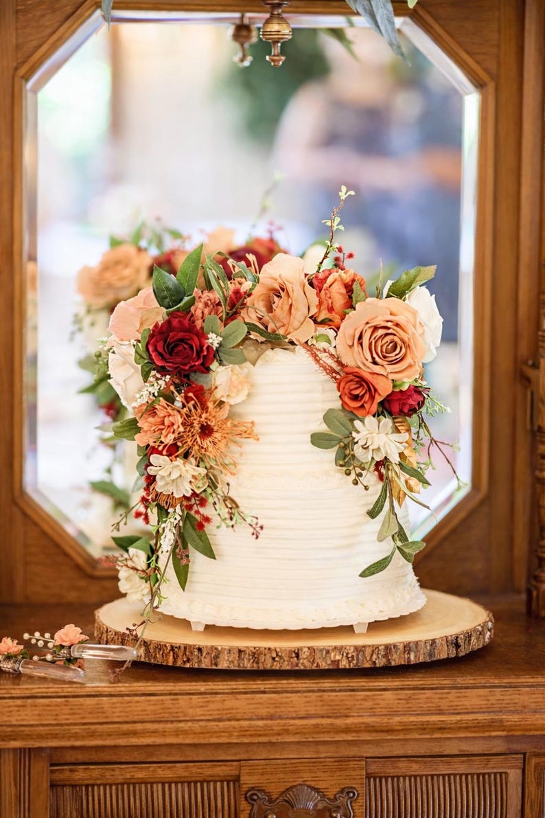 Puede incluir: Un pastel blanco de dos niveles con glaseado de textura horizontal, decorado con un arreglo de rosas artificiales y follaje en tonos rojo intenso, naranja quemado, melocot&oacute;n y crema, sobre una base de madera natural.