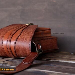 May include: A brown leather satchel bag with a flap closure and a strap. The bag is on a wooden surface with a small wooden tool nearby. The text "LEATHERCRAFT" is visible on the bag.