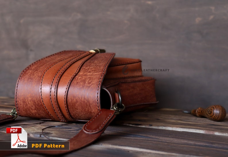 May include: A brown leather satchel bag with a flap closure and a strap. The bag is on a wooden surface with a small wooden tool nearby. The text "LEATHERCRAFT" is visible on the bag.