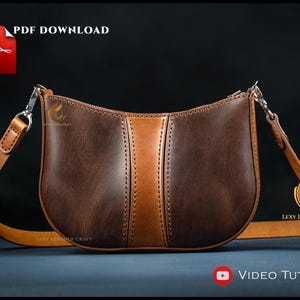 May include: A brown leather shoulder bag with a lighter brown vertical stripe down the center. The bag has a curved shape and a long, adjustable strap. The words "PDF DOWNLOAD" and "VIDEO TUTORIAL" are visible.