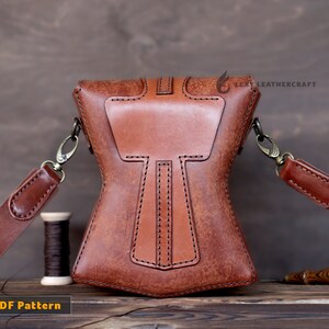 May include: Brown leather crossbody bag with a unique design featuring a large, central, rectangular flap with a smaller, rectangular flap on top. The bag has a long, adjustable strap and a small, decorative metal clasp.