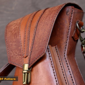 May include: A brown leather bag with a flap closure and a metal buckle. The bag has a shoulder strap and is stitched with brown thread. The bag has the text "Lexy Leathercraft" on the side.