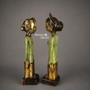 May include: Two tall standing figurines facing each other, featuring polished gold-tone heads and bases with a textured green patina on the torso sections. The text Borneo Hunters is centred between the figures. Each sculpture has a distinct hairstyle.