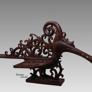 May include: A dark brown wooden sculpture of a bird with intricate carvings. The bird has a long beak and its wings are spread out. The sculpture is on a wooden base and has the text "Borneo Hunters" on the front.