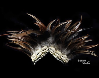 May include: A brown and black feather headdress with white seashells and gold beads. The headdress is on a black background. Borneo Hunters.
