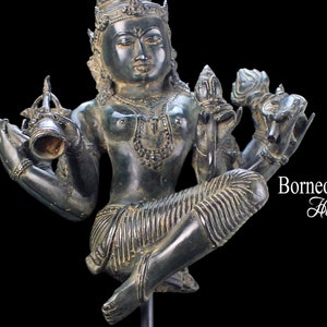 Puede incluir: Una estatua de bronce verde oscuro de una deidad hindú con cuatro brazos, sosteniendo una concha, una flor de loto y una campana. La estatua está sentada sobre una base negra.