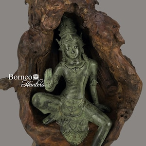 Puede incluir: Una estatua de bronce verde de una deidad sentada dentro de un trozo de madera ahuecado. La madera es de color marrón oscuro y tiene una textura rugosa. La estatua está sobre una base negra con un marco de madera.