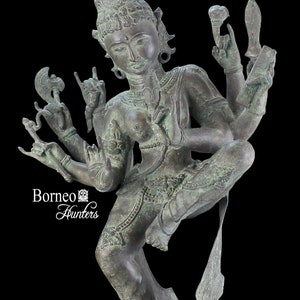 Puede incluir: Una estatua de bronce de una deidad hindú con múltiples brazos, que lleva una corona y joyas elaboradas, de pie sobre una base de madera de varios niveles. La estatua está en una pose de baile, con una pierna levantada y la otra doblada. La deidad tiene una expresión serena en su rostro.