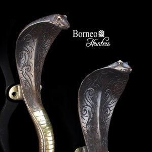 May include: Two decorative door handles in the shape of cobra snakes. The handles are made of metal with a dark finish and brass accents. The cobra heads are detailed with etched patterns. The handles are mounted on brass plates.