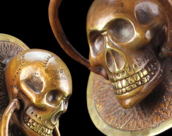 May include: Two brass skull door knockers with intricate details. Each knocker features a skull with a toothy grin and a ring for knocking. The knockers are mounted on circular plates with decorative patterns.