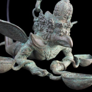 May include: A green bronze sculpture of a mythical bird-like creature with wings outstretched, holding three small bowls. The sculpture is hanging from a chain.
