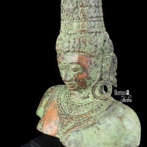 Puede incluir: Un busto de bronce verde de una figura femenina que lleva una corona y joyas ornamentadas. El busto está sobre una base de madera negra. Borneo Hunters está impreso en la base.