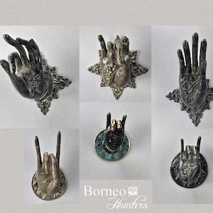 Puede incluir: Colección de seis objetos decorativos antiguos con forma de mano en tono plateado. Cada mano está en un gesto diferente, montada sobre bases ornamentadas en forma de estrella o circulares. El logotipo de Borneo Hunters es visible en la esquina inferior derecha.