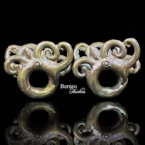 May include: Two ornate bronze rings with intricate swirling designs. The rings are connected by a small, round, decorative piece. The rings are on a black background.
