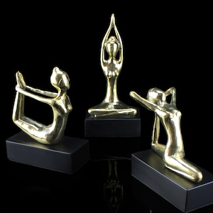 May include: Three gold-toned metal sculptures of people in yoga poses on black bases. The sculptures are abstract and stylized.
