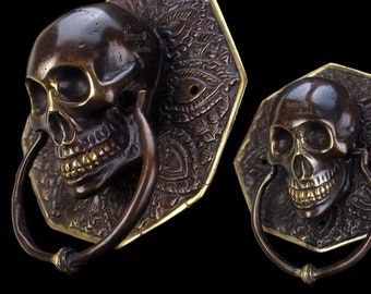 May include: Two ornate brass door knockers shaped like skulls with intricate details. Each knocker has a ring for pulling and is mounted on an octagonal base with a decorative pattern.