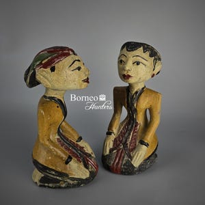 Puede incluir: Dos figuras de madera talladas a mano con vestimenta tradicional. Las figuras están pintadas con detalles en amarillo, rojo y negro. Una figura lleva un tocado con detalles verdes y rojos. Las figuras están arrodilladas con las manos en las rodillas.