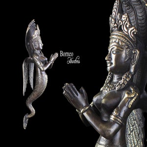 May include: Two bronze statues of a female figure with wings and a snake tail. The statues are in a praying position with their hands clasped together. The statues are both facing the right side of the image. The statue on the left is smaller than the statue on the right. The statue on the right has a large headdress with intricate details. The text "Borneo Hunters" is visible in the background.