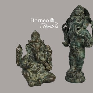 Puede incluir: Dos estatuas de bronce del dios hindú Ganesha. La estatua de la izquierda está sentada con la trompa enroscada, mientras que la estatua de la derecha está de pie con la trompa apuntando hacia adelante. Ambas estatuas tienen detalles intrincados y una pátina verde.