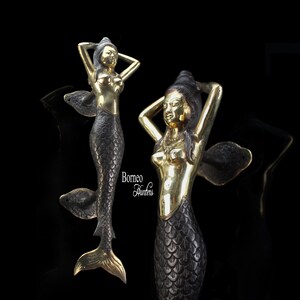 Könnte beinhalten: Zwei goldfarbene Meerjungfrau-Türklopfer mit schwarzen und goldenen Details. Die Meerjungfrauen sind mit langen, fließenden Haaren dargestellt und halten sich die Haare in den Händen. Die Meerjungfrauen haben einen schwarzen und goldenen Fischschwanz.