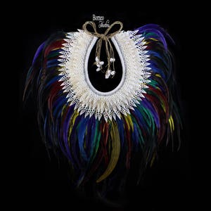 Puede incluir: Un collar de conchas blancas con un diseño de encaje y un borde de plumas multicolores. El collar está atado con un cordón marrón y tiene cuentas blancas colgando de él. El texto "Borneo Hunters" es visible en el cordón.