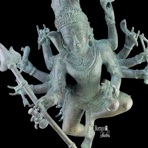 Puede incluir: Estatua de color bronce de una deidad de múltiples brazos, posiblemente hindú, con detalles intrincados. La figura está adornada con un tocado detallado y sostiene varios objetos. La estatua tiene una apariencia desgastada, lo que sugiere edad o un acabado específico. El fondo es negro.