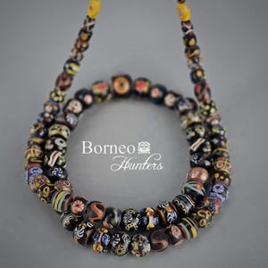Puede incluir: Un collar con una cadena de cuentas coloridas y estampadas. Las cuentas son principalmente negras con diseños intrincados en varios colores, incluyendo amarillo, rojo y azul. El texto "Borneo Hunters" es visible.