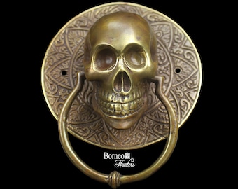 May include: A brass door knocker shaped like a skull with intricate details. The skull is surrounded by a circular design with a ring handle.
