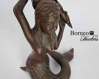 May include: Bronze mermaid sculpture holding a seashell. The mermaid has long hair and a scaled tail. The sculpture has a weathered, antique finish. The text "Borneo Hunters" is visible in the image.