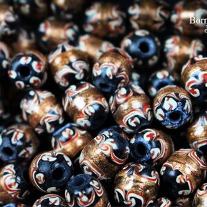 May include: A close-up of a collection of decorative beads. The beads are made of a dark blue material with white and red swirls and a gold metallic finish. The beads are arranged in a random pattern.