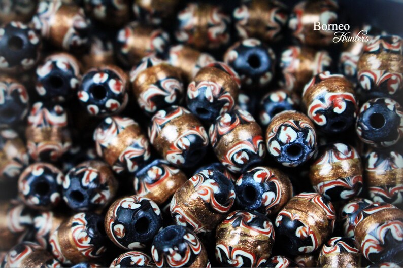 May include: A close-up of a collection of decorative beads. The beads are made of a dark blue material with white and red swirls and a gold metallic finish. The beads are arranged in a random pattern.