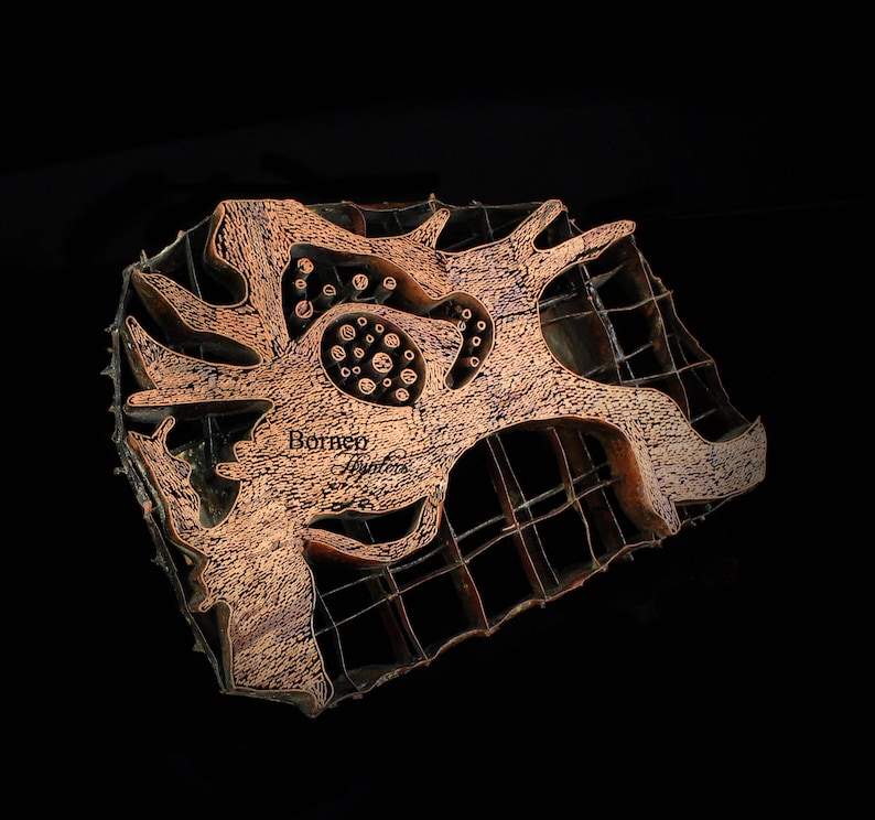 May include: A copper-colored batik stamp with a complex design featuring a central motif of a stylized flower with a grid pattern surrounding it. The stamp is labeled "Borneo Batiks".
