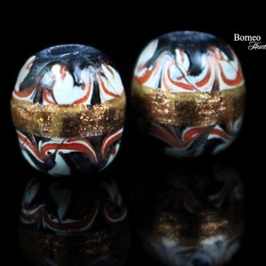 May include: Two round glass beads with a white, black, and red marbled pattern. Each bead has a gold band around the middle.