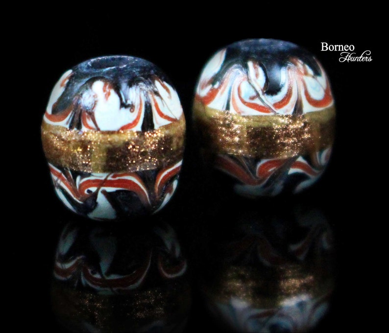 May include: Two round glass beads with a white, black, and red marbled pattern. Each bead has a gold band around the middle.