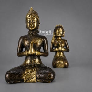 Peut inclure: Deux statues en bronze représentant des personnages en position de prière assise, les mains jointes. Les figurines présentent des motifs floraux en relief sur leurs vêtements et coiffes. Le texte Borneo Hunters est visible au centre.