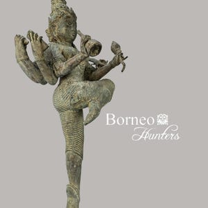 Puede incluir: Una estatua de bronce de una deidad hindú de múltiples brazos, posiblemente Shiva, con una pátina verde. La deidad está de pie con una pierna levantada y sosteniendo varios objetos en sus manos. La estatua está sobre una base negra. Borneo Hunters.