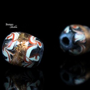 May include: Two colorful glass beads with intricate swirling patterns in shades of red, white, black, and gold. The beads have a slightly irregular shape and a glossy finish.