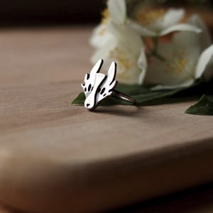 Könnte beinhalten: Ein silberner Ring mit einem stilisierten Drachenkopf-Design. Der Ring hat ein detailliertes, durchbrochenes Muster mit einem Drachengesicht und Flügeln. Der Ring wird auf einer Holzoberfläche mit weißen Blumen im Hintergrund präsentiert.
