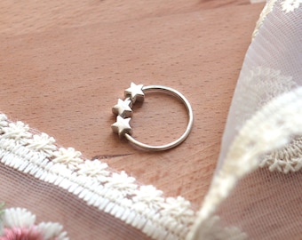 May include: A silver ring with three star-shaped accents. The ring is on a wooden surface with a white lace fabric in the background.