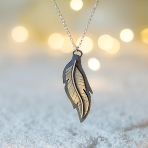May include: A silver-toned feather pendant necklace. The pendant features a detailed feather design with a contrasting gold-colored interior. The necklace hangs from a delicate silver chain. The background is blurred with soft, warm bokeh lights.