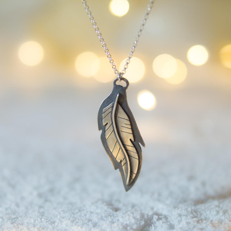 May include: A silver-toned feather pendant necklace. The pendant features a detailed feather design with a contrasting gold-colored interior. The necklace hangs from a delicate silver chain. The background is blurred with soft, warm bokeh lights.