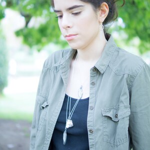 May include: A silver necklace with two pendants, one a leaf shape and the other a textured geometric shape, hangs over a black top. The model is wearing a green button-down shirt and silver hoop earrings. The background is blurred with green foliage.